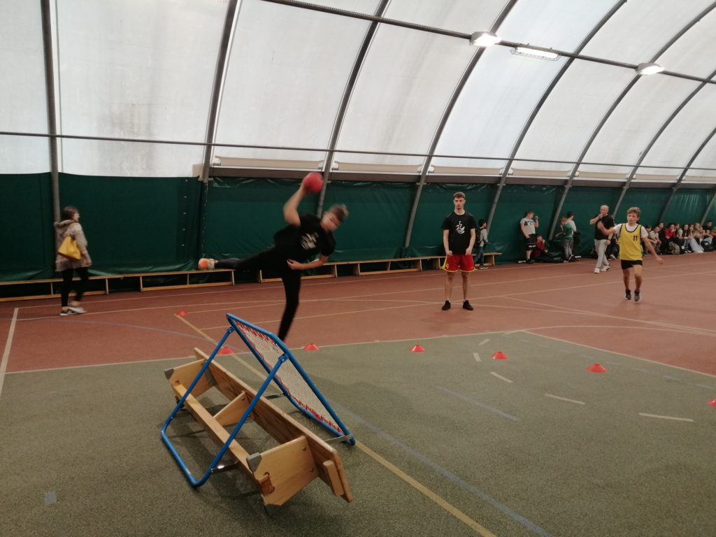 Zdjęcie: mecz pokazowy, uczniowie na boisku, zdjęcie grupowe. Zawodnik Tchoukball Lubuskie wykonuje rzut z wyskoku na bramkę. 